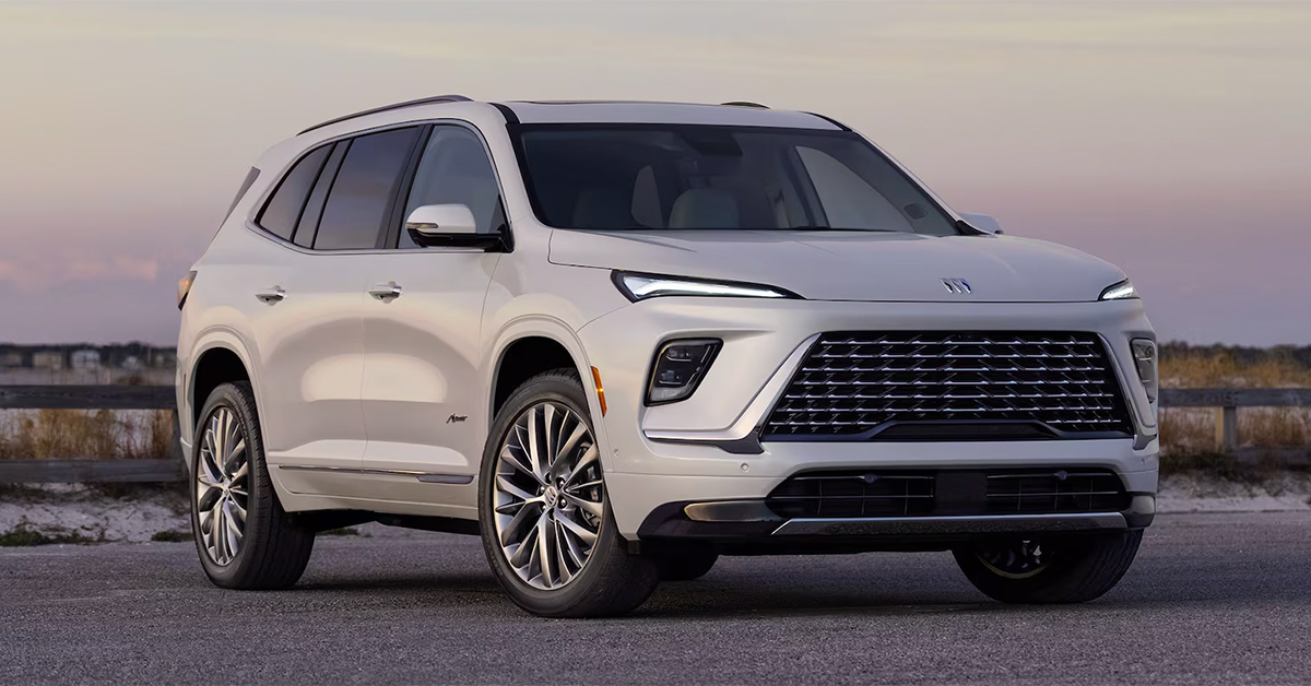 A sleek white SUV is parked on a paved road with a sunset sky in the background. It has modern headlights and a large grille, conveying elegance and power.